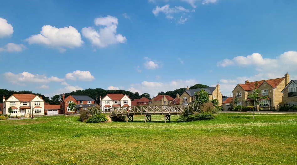 Houses in the background with a communal green area in the foreground. A wooden structure is in the middle of the green area.