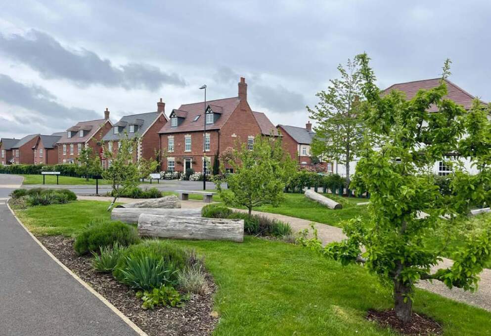 Houses in the background with a communal green space including benches, logs and trees.