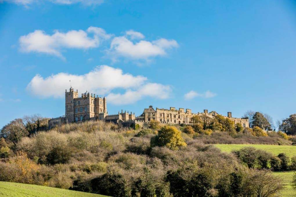 A medieval castle with multiple towers and battlements atop a hill, surrounded by autumn trees and green fields under a blue sky with scattered white clouds.