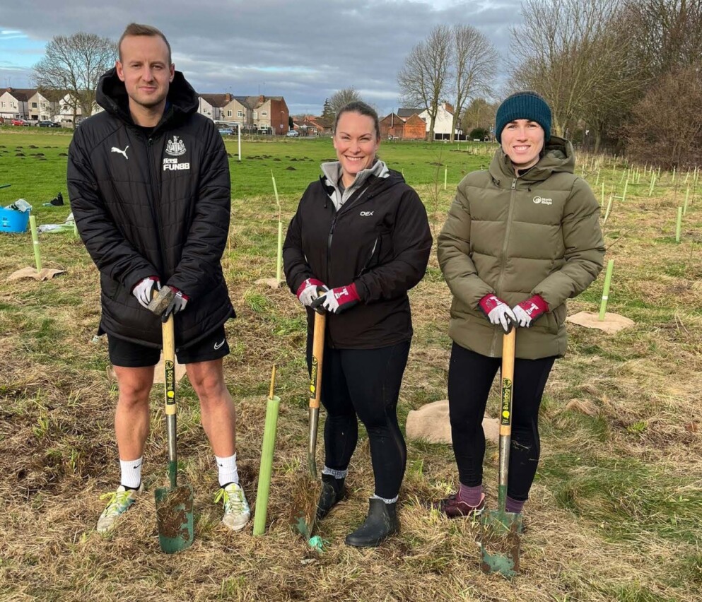 Three people standing outdoors on a grassy field with small trees marked by green stakes around them, holding shovels with yellow handles. They wear casual clothing and gloves, with houses and leafless trees in the background under a cloudy sky.