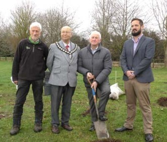 Four individuals standing outdoors on grass, with leafless trees in the background. The second person from the left wears a gray blazer with a decorative collar, the third holds a shovel with a painted handle, and the fourth is in a dark blazer and khakis.