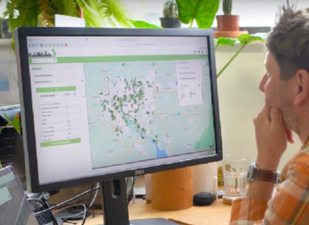 Blurry profile of a person wearing an orange plaid shirt, sitting at a desk with a water glass, looking at a computer screen displaying a map interface with numerous green markers.